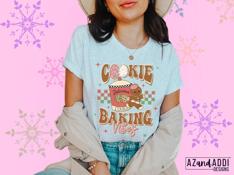 May include: Light blue t-shirt with the words "COOKIE BAKING VIBES" in brown and green retro font. The graphic includes a cookie jar, gingerbread man, and cookies. The shirt is worn by a person.