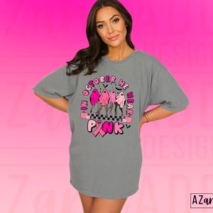 May include: Gray t-shirt with a graphic that reads "In October We Wear Pink." The design features three ghosts in pink outfits, a pink ribbon, and Halloween-themed elements. The shirt is a comfortable, oversized fit.