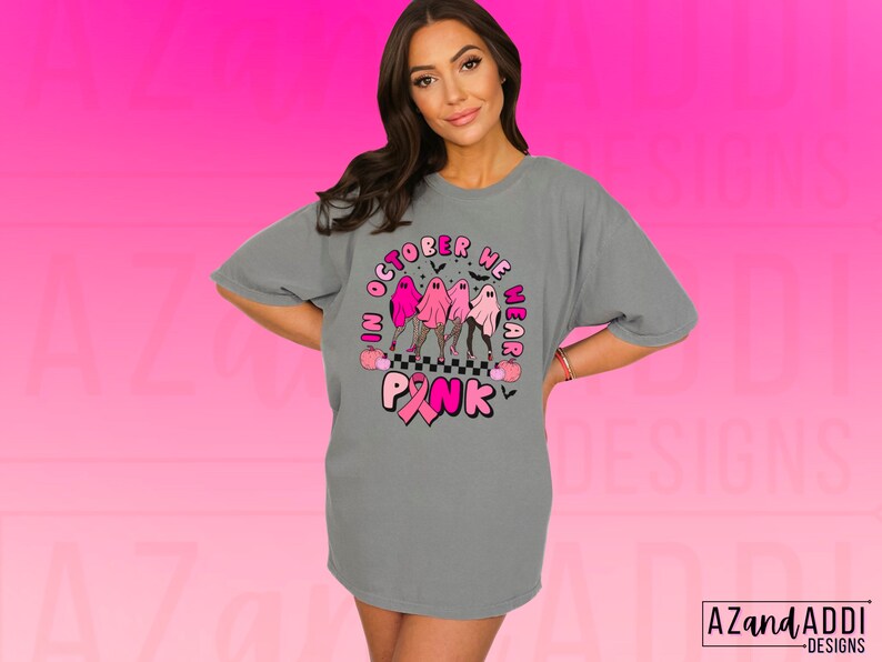 May include: Gray t-shirt with a graphic that reads "In October We Wear Pink." The design features three ghosts in pink outfits, a pink ribbon, and Halloween-themed elements. The shirt is a comfortable, oversized fit.
