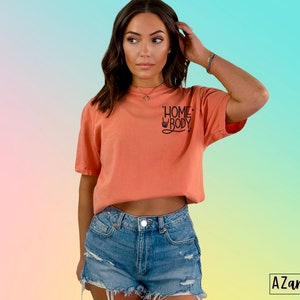 Puede incluir: Una camiseta de color melocot&oacute;n con el texto "Homebody" en letras negras. La camiseta la lleva puesta una persona con el pelo largo y casta&ntilde;o y se combina con unos pantalones cortos de mezclilla.