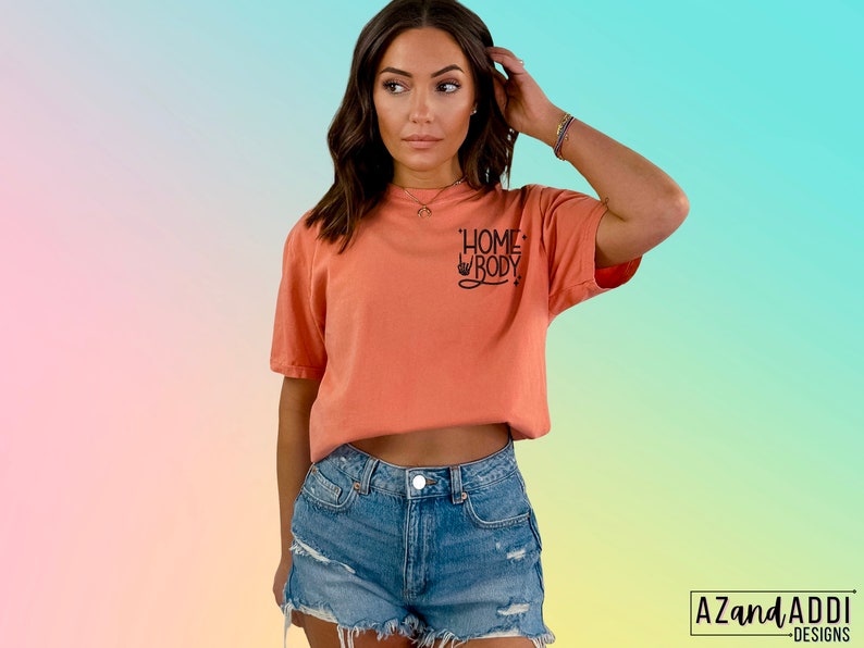 Puede incluir: Una camiseta de color melocot&oacute;n con el texto "Homebody" en letras negras. La camiseta la lleva puesta una persona con el pelo largo y casta&ntilde;o y se combina con unos pantalones cortos de mezclilla.