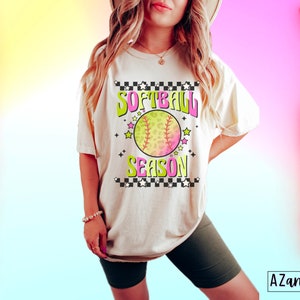 May include: A white t-shirt with a pink, green, and yellow graphic that says "Softball Season" with a softball graphic. The shirt is being worn by a person with long blonde hair. The background is a gradient of pink, yellow, and blue.