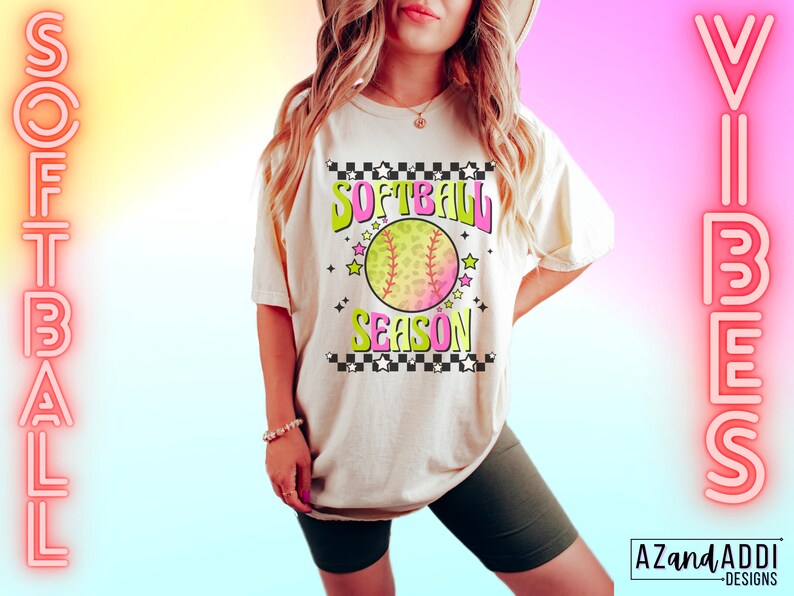 May include: A white t-shirt with a pink, green, and yellow graphic that says "Softball Season" with a softball graphic. The shirt is being worn by a person with long blonde hair. The background is a gradient of pink, yellow, and blue.