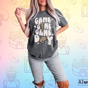 May include: A gray t-shirt with the text "Game Game Game Day" in a retro font. The shirt features a lightning bolt and a football helmet with a leopard print design.