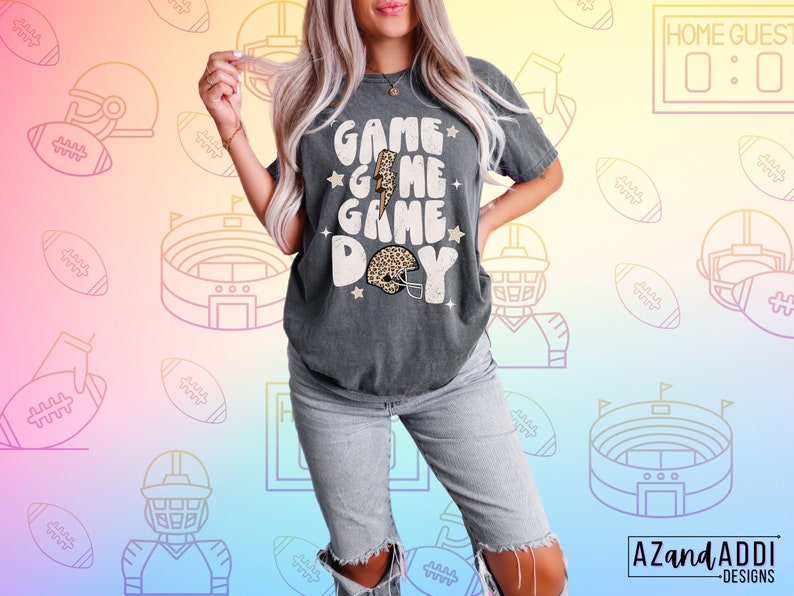 May include: A gray t-shirt with the text "Game Game Game Day" in a retro font. The shirt features a lightning bolt and a football helmet with a leopard print design.