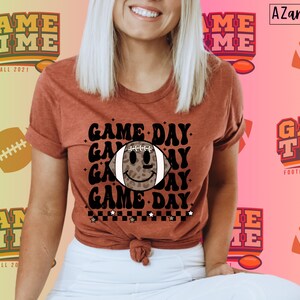 May include: A rust-coloured t-shirt with the words "GAME DAY" and a football graphic with a smiley face. The shirt also features the text "GAME TIME FOOTBALL 2021" and a football design. The shirt is a short-sleeved tee.