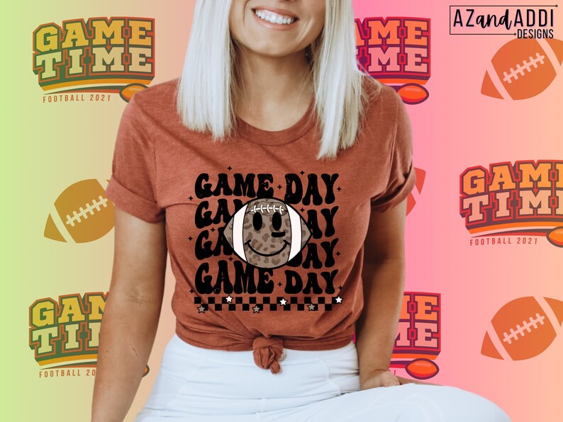 May include: A rust-coloured t-shirt with the words "GAME DAY" and a football graphic with a smiley face. The shirt also features the text "GAME TIME FOOTBALL 2021" and a football design. The shirt is a short-sleeved tee.