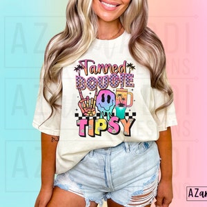 May include: A white t-shirt featuring a colourful graphic design that reads "Tanned Bougie Tipsy". The graphic includes a smiley face wearing sunglasses, a palm tree, and a drink.