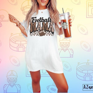 Pode incluir: Camiseta branca com texto marrom que diz "Football Mama" com um gr&aacute;fico de futebol e um padr&atilde;o xadrez. A camiseta &eacute; de tamanho grande e tem um caimento solto.