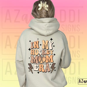 May include: A white hoodie with a graphic on the back that says "In My Hockey Mom Era" in a retro font with a hockey stick and puck design.