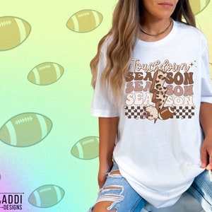 May include: White t-shirt with a brown and white graphic that says "Touchdown Season" with a lightning bolt and football design.