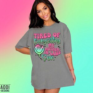 Puede incluir: Camiseta gris con un gr&aacute;fico rosa y azul que dice "Cansado de cuidar al hijo de mi suegra".