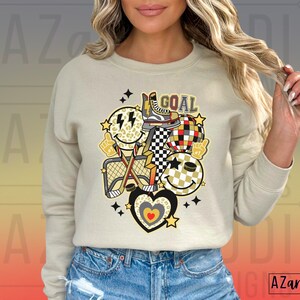 May include: A beige sweatshirt with a graphic design of a hockey stick, a net, a puck, and smiley faces. The graphic also includes the word "GOAL" in black letters.