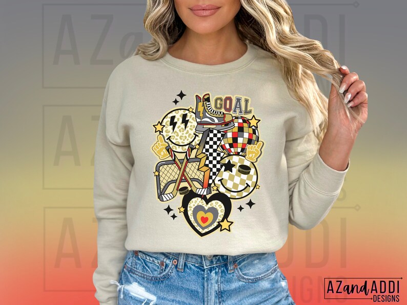 May include: A beige sweatshirt with a graphic design of a hockey stick, a net, a puck, and smiley faces. The graphic also includes the word "GOAL" in black letters.
