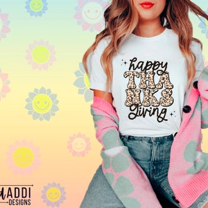 May include: White t-shirt with the text "happy thanks giving" in black with a leopard print design. The shirt is being worn by a person with a pink cardigan.