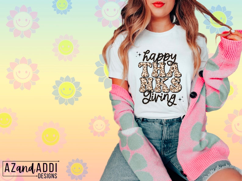May include: White t-shirt with the text "happy thanks giving" in black with a leopard print design. The shirt is being worn by a person with a pink cardigan.