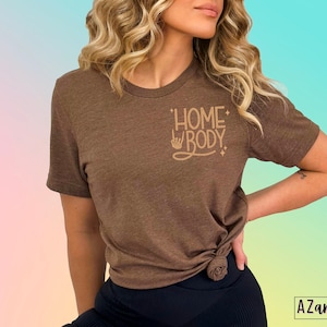 Puede incluir: Una camiseta marr&oacute;n con el texto "Homebody" en letras doradas. El texto est&aacute; estilizado con un signo de paz y estrellas.