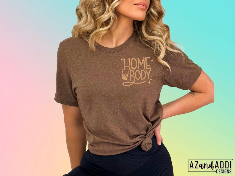 Puede incluir: Una camiseta marr&oacute;n con el texto "Homebody" en letras doradas. El texto est&aacute; estilizado con un signo de paz y estrellas.