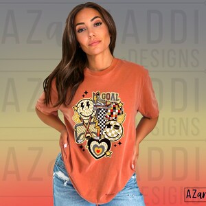May include: A rust-colored t-shirt with a graphic design featuring a smiley face, a checkered pattern, a heart, and the word "GOAL".
