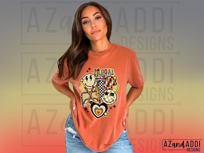 May include: A rust-colored t-shirt with a graphic design featuring a smiley face, a checkered pattern, a heart, and the word "GOAL".