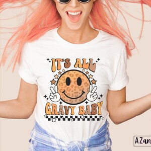 May include: White t-shirt with a smiling face graphic with a leopard print design. The text "It's All Gravy Baby" is printed on the shirt.
