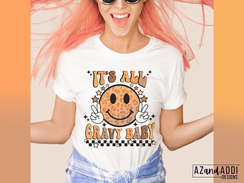 May include: White t-shirt with a smiling face graphic with a leopard print design. The text "It's All Gravy Baby" is printed on the shirt.