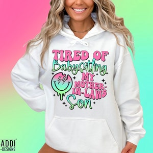 Puede incluir: Sudadera blanca con un gr&aacute;fico rosa, verde y azul que dice "Cansado de cuidar al hijo de mi suegra".