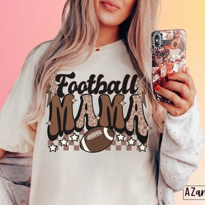 Pode incluir: Uma camiseta branca com um design gr&aacute;fico marrom e preto que diz "Football Mama" com uma bola de futebol e estrelas.