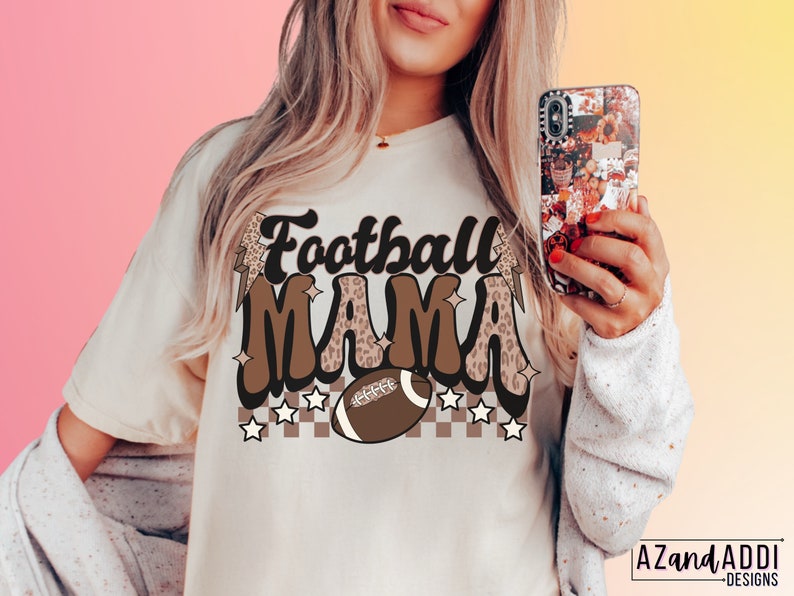 Pode incluir: Uma camiseta branca com um design gr&aacute;fico marrom e preto que diz "Football Mama" com uma bola de futebol e estrelas.