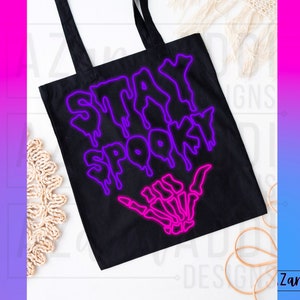 May include: Black tote bag with a neon pink and purple graphic that says "Stay Spooky" with a skeleton hand making a shaka sign.