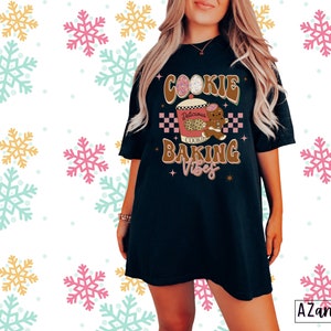 May include: Black t-shirt with the text "Cookie Baking Vibes" in brown and pink. The graphic includes a gingerbread man, a cookie, and a coffee cup. The shirt has a relaxed fit and short sleeves. The background has a snowflake pattern.