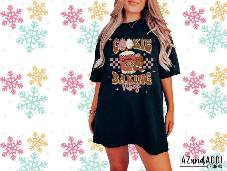May include: Black t-shirt with the text "Cookie Baking Vibes" in brown and pink. The graphic includes a gingerbread man, a cookie, and a coffee cup. The shirt has a relaxed fit and short sleeves. The background has a snowflake pattern.