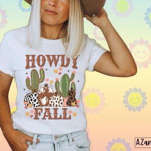 May include: White t-shirt with a graphic design featuring the text "HOWDY FALL" in brown, along with illustrations of cacti and pumpkins. The pumpkins have various patterns, including cow print and leopard print.
