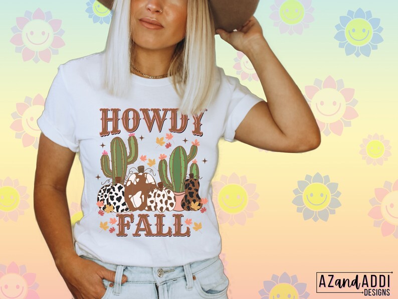 May include: White t-shirt with a graphic design featuring the text "HOWDY FALL" in brown, along with illustrations of cacti and pumpkins. The pumpkins have various patterns, including cow print and leopard print.