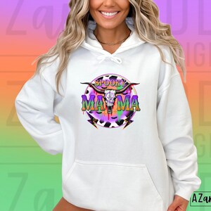May include: White hoodie with a colourful graphic. The design includes a cow skull, the words "Spooky Mama", and lightning bolts. The graphic uses purple, green, and orange, with a cow print background. The hoodie has a drawstring hood and a front pocket.