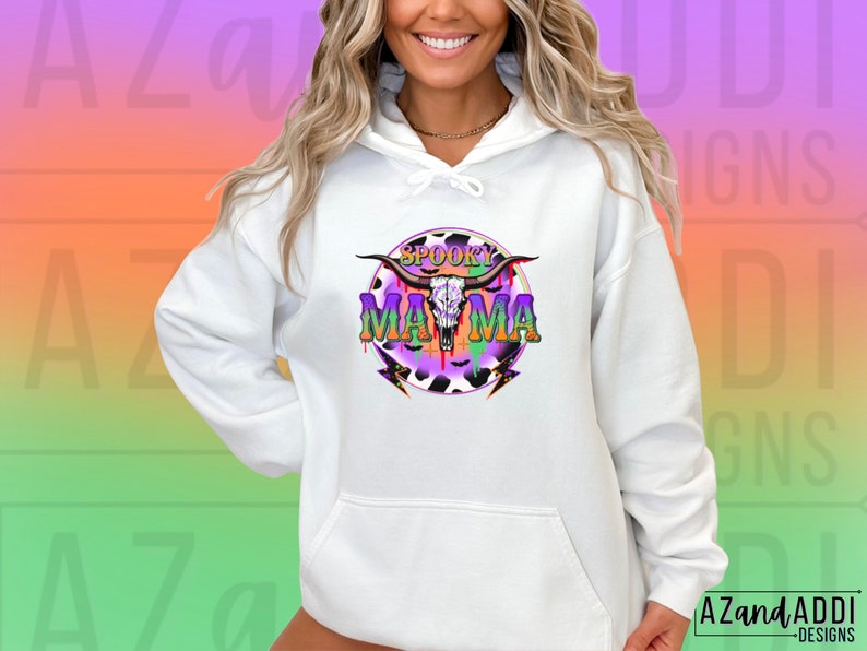 May include: White hoodie with a colourful graphic. The design includes a cow skull, the words "Spooky Mama", and lightning bolts. The graphic uses purple, green, and orange, with a cow print background. The hoodie has a drawstring hood and a front pocket.