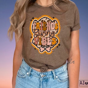May include: A brown t-shirt with the words "FRIENDS GIVING VIBES" in a retro font. The graphic has a leopard print design with orange, pink, and white accents. The shirt is worn by a person wearing blue jeans.