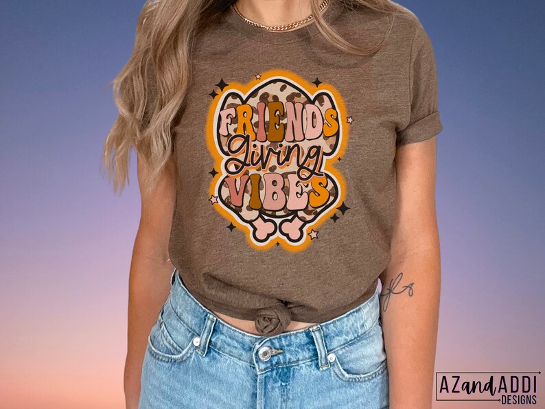 May include: A brown t-shirt with the words "FRIENDS GIVING VIBES" in a retro font. The graphic has a leopard print design with orange, pink, and white accents. The shirt is worn by a person wearing blue jeans.