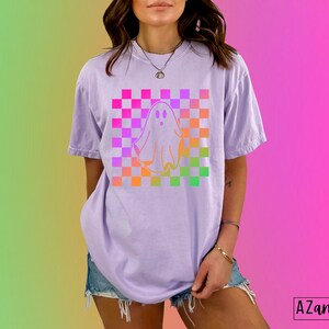 Op de afbeelding: Lichtpaars T-shirt met een spookontwerp op een geruite achtergrond in roze, oranje en groen. Het shirt heeft korte mouwen en een relaxte pasvorm. De tekst "AZandADDI DESIGNS" staat rechtsonder.