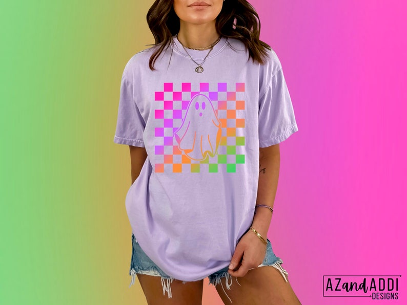 Op de afbeelding: Lichtpaars T-shirt met een spookontwerp op een geruite achtergrond in roze, oranje en groen. Het shirt heeft korte mouwen en een relaxte pasvorm. De tekst "AZandADDI DESIGNS" staat rechtsonder.