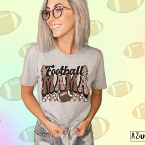 Pode incluir: Uma camiseta cinza com o texto "Football Mama" em letras marrons e pretas com um design de estampa de leopardo. O texto &eacute; cercado por estrelas e uma bola de futebol.