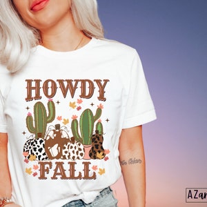 May include: White t-shirt with a fall-themed graphic. The design features the text "HOWDY FALL" in brown letters, along with illustrations of cacti, pumpkins, and autumn leaves. The shirt is a crew neck style.