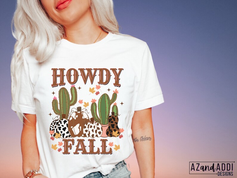 May include: White t-shirt with a fall-themed graphic. The design features the text "HOWDY FALL" in brown letters, along with illustrations of cacti, pumpkins, and autumn leaves. The shirt is a crew neck style.