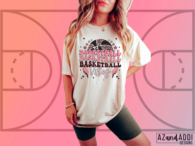 May include: Cream-colored t-shirt with a basketball-themed graphic. The design features the words "BASKETBALL VIBES" in pink and black, along with a basketball and lightning bolt. The shirt is paired with dark green shorts.