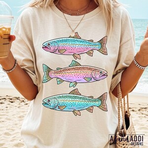 Puede incluir: Camiseta beige con tres ilustraciones de peces coloridos. Cada pez presenta una combinaci&oacute;n de colores &uacute;nica, incluyendo azul, rosa y verde, con dise&ntilde;os detallados de aletas y escamas. La camiseta tiene un corte relajado y es adecuada para uso casual.