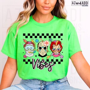 May include: A neon green t-shirt featuring a graphic with three cartoon characters: a scuba diver, Jason Voorhees, and Chucky. The word "Vibes" is written in a cursive font below the characters. The design includes black and white checkered patterns and seashell accents.