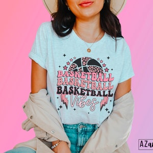 May include: Light gray t-shirt with a basketball graphic. The design features a basketball with a leopard print pattern, stars, and the words "Basketball Vibes" in pink and black. The shirt has rolled-up sleeves.