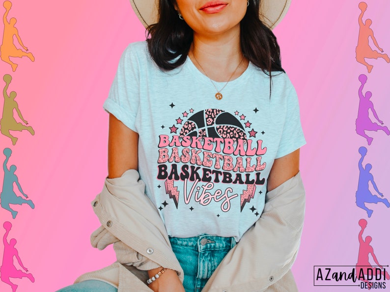 May include: Light gray t-shirt with a basketball graphic. The design features a basketball with a leopard print pattern, stars, and the words "Basketball Vibes" in pink and black. The shirt has rolled-up sleeves.
