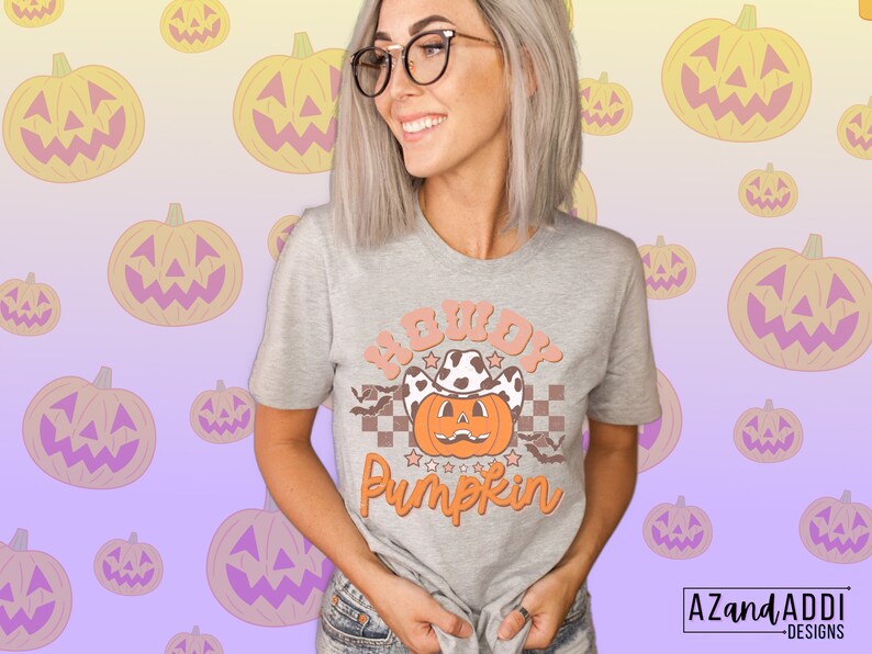 May include: A light gray t-shirt with the text "Howdy Pumpkin" in orange and peach colors. The design features a pumpkin wearing a cowboy hat and stars. The shirt is a Halloween-themed graphic tee.
