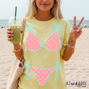 K&ouml;nnte beinhalten: Hellgelbes T-Shirt mit Bikini-Design. Das Bikini-Oberteil und -Unterteil sind mit rosa und orangefarbenen Karos versehen, die hellblau umrandet sind. Das T-Shirt hat einen l&auml;ssigen Stil, ideal f&uuml;r einen Tag am Strand. Das Design enth&auml;lt den Text "AZandADDI DESIGNS".